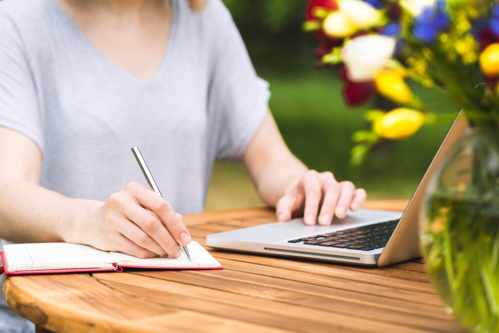 Hands writing planning notes in a notebook beside a laptop